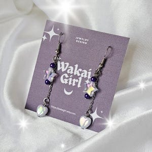 Könnte beinhalten: Ohrringe zum Aufhängen mit silberfarbenen Haken, sternförmigen Anhängern, lila Perlen und herzförmigen Anhängern. Die Ohrringe werden auf einer lila Karte mit dem Text "Wakai Girl" und "Jewelry Design" präsentiert.