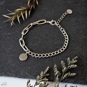 Könnte beinhalten: Silberfarbenes Armband mit gemischtem Ketten-Design. Ein Abschnitt hat längliche Glieder, der andere ein Panzerketten-Design. Ein kleiner, runder Anhänger hängt am Abschnitt mit den länglichen Gliedern. Das Armband wird auf einer dunklen Oberfläche mit getrocknetem Blattwerk präsentiert.