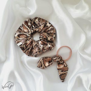 Op de afbeelding: Twee haaraccessoires: een grote scrunchie en een haarband met strik, beide met een slangenprint in bruin, beige en wit. De scrunchie is gerimpeld en de haarband heeft een roze elastiek.