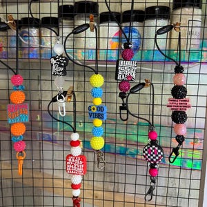 Puede incluir: Una colección de cordones con cuentas con varios diseños y texto. Los cordones presentan cuentas coloridas, dijes y frases como "Good Vibes" y "Bad Bitch Have Bad Days 100". Cada cordón tiene un clip de metal.
