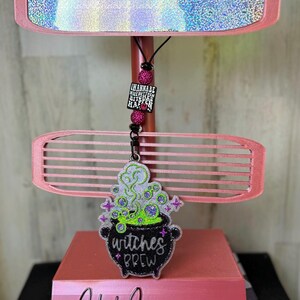 May include: A pink display stand with a holographic top and a decorative, pink, horizontal slat. A charm with the text "I wannabe nice but then bitches happen" hangs from the top. A cauldron charm with "witches brew" is also present.