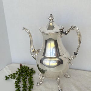May include: A polished silver coffee pot with a decorative handle and spout. The pot has a lid with a finial and rests on three ornate feet. The pot is reflective, and the background is a neutral color.