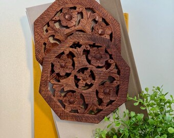 May include: Two intricately carved wooden coasters with a floral design. The coasters are brown and have a natural wood finish.