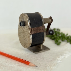 May include: A vintage manual pencil sharpener with a dark black cylindrical body and a weathered, speckled metallic front plate. It features a side-mounted hand crank with a small knob and a flat metal base. An orange pencil lies in the foreground.