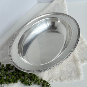 May include: A silver oval serving platter with a decorative border. The platter is placed on a white linen napkin with a fringed edge. Green leafy sprigs are in the lower left corner.