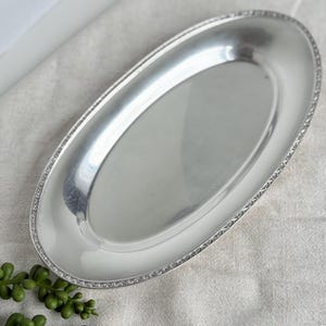 May include: A silver-colored, oval serving tray with a decorative beaded edge. The tray is reflective and sits on a neutral-colored surface. The tray is empty and ready for use.