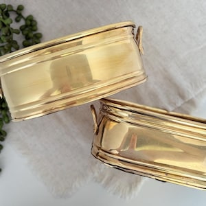 May include: Two rectangular, gold-colored brass planters with small handles. The planters have a shiny finish and are placed on a white cloth. Greenery is visible in the background. These planters are suitable for indoor or outdoor use.