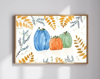 May include: Watercolor painting of three pumpkins in shades of blue, orange, and green. The pumpkins are arranged on a white background and framed by autumn leaves in gold and blue. The artwork is in a wooden frame.
