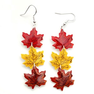 May include: A pair of dangle earrings featuring three layered maple leaves. The leaves are red, orange, and yellow with black veins and are made of resin. The earrings have silver hooks.