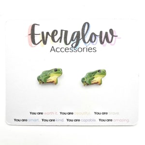 May include: A pair of frog-shaped earrings on a white card. The earrings are green, yellow, and brown. The card has the text "Everglow Accessories" and inspirational phrases like "You are worth it."