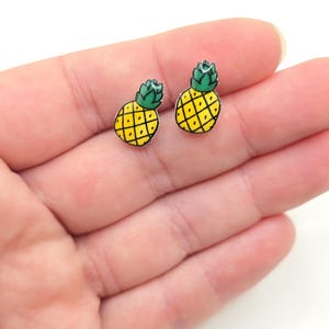 May include: A pair of silver stud earrings featuring a yellow pineapple with a green top. The earrings are shown on a hand.