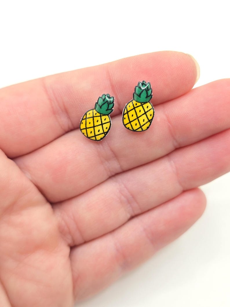 May include: A pair of silver stud earrings featuring a yellow pineapple with a green top. The earrings are shown on a hand.