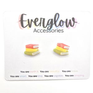 May include: A pair of book-shaped stud earrings on a white card. The earrings are miniature stacks of books with red, yellow, and green covers. The card has the text "Everglow Accessories" and inspirational phrases.