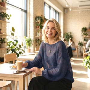 Pode incluir: Uma mulher sorri num café, segurando um café. Ela usa um suéter azul e um colar. O café é luminoso com plantas e luz natural, criando uma atmosfera relaxante.