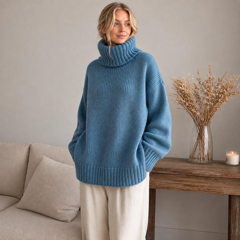 May include: A chunky knit sweater in a dusty blue hue featuring a prominent ribbed turtleneck collar, long sleeves with ribbed cuffs, and a wide ribbed hemline. The garment is crafted with a thick, textured knit pattern throughout the body.