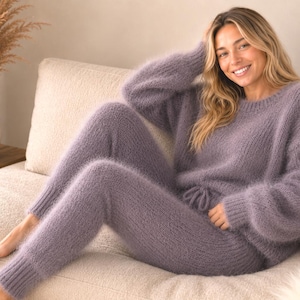 May include: A matching two-piece lounge set in a soft, fuzzy, muted purple knit. The set consists of a long-sleeved jumper with a round neckline and ribbed cuffs, paired with matching high-waisted knit trousers featuring a drawstring waistband.