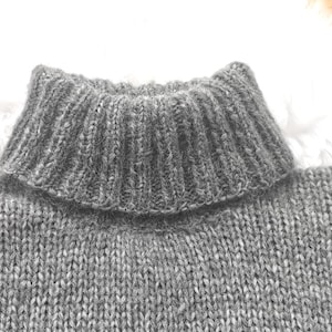 May include: Close-up of a gray knit sweater with a ribbed turtleneck collar. The sweater has a textured appearance, with visible knit stitches. The collar is folded over, adding a layered effect to the design. The overall style suggests a warm, comfortable garment.