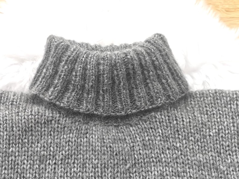 May include: Close-up of a gray knit sweater with a ribbed turtleneck collar. The sweater has a textured appearance, with visible knit stitches. The collar is folded over, adding a layered effect to the design. The overall style suggests a warm, comfortable garment.