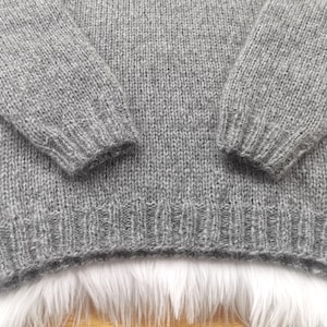 May include: Close-up of a gray knit sweater. The sweater has long sleeves and a ribbed hem and cuffs. The knit fabric has a textured appearance. The sweater is made of a soft, warm material.