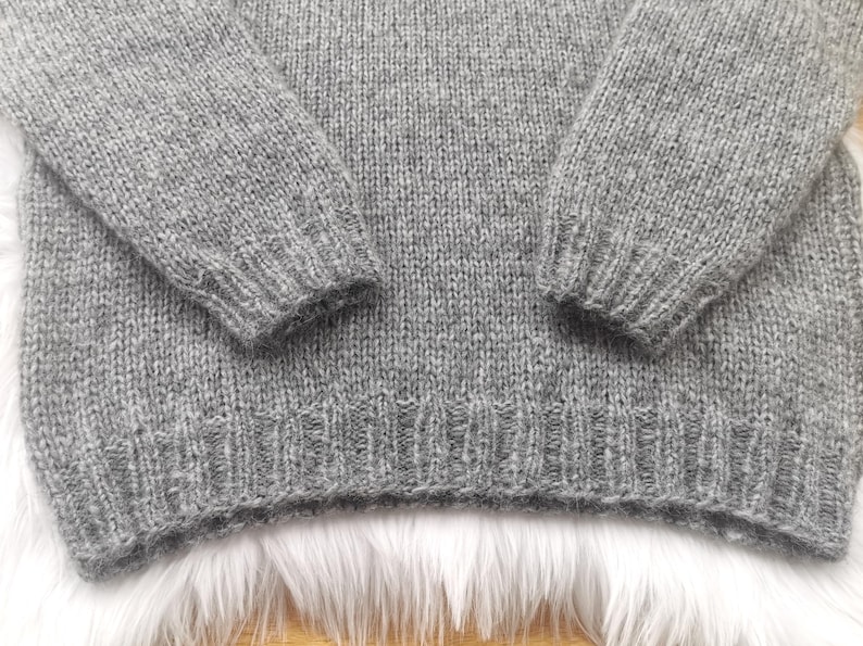 May include: Close-up of a gray knit sweater. The sweater has long sleeves and a ribbed hem and cuffs. The knit fabric has a textured appearance. The sweater is made of a soft, warm material.