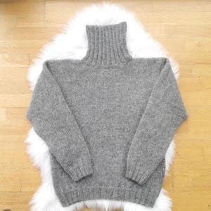 May include: A gray turtleneck sweater with long sleeves. The sweater is made of a knitted material and is displayed on a white fluffy surface, set against a light brown wooden background. The sweater has a ribbed collar and cuffs.