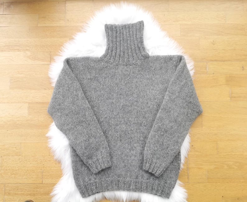 May include: A gray turtleneck sweater with long sleeves. The sweater is made of a knitted material and is displayed on a white fluffy surface, set against a light brown wooden background. The sweater has a ribbed collar and cuffs.