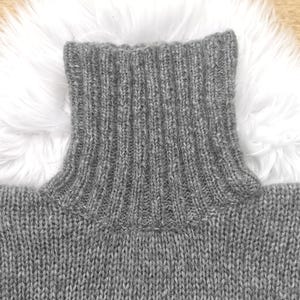 May include: Close-up of a gray turtleneck sweater. The ribbed collar is prominent, and the knit fabric has a textured appearance. The sweater is made of a soft, warm material, perfect for cold weather. The background is a white fluffy surface.