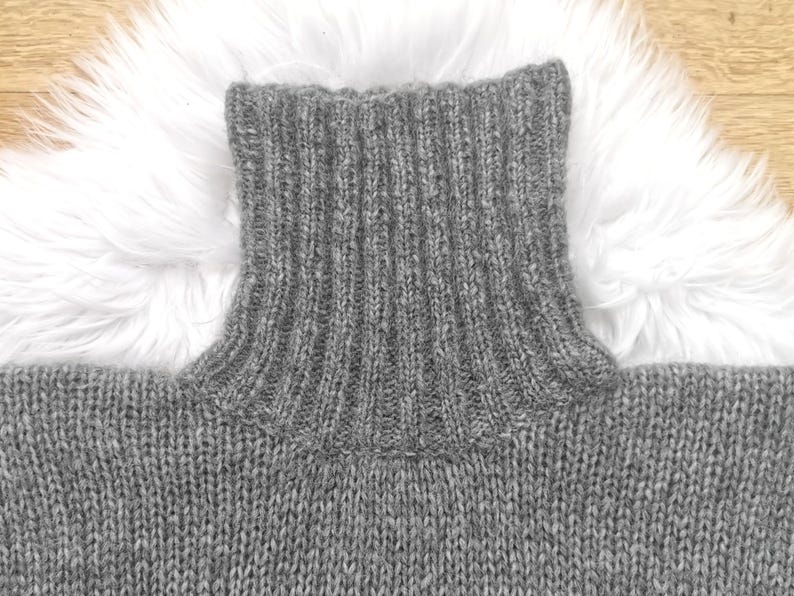 May include: Close-up of a gray turtleneck sweater. The ribbed collar is prominent, and the knit fabric has a textured appearance. The sweater is made of a soft, warm material, perfect for cold weather. The background is a white fluffy surface.