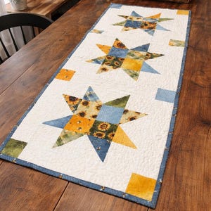 May include: A quilted table runner with a white background and a repeating star pattern. The stars are made of blue, yellow, and green fabric with sunflower and bee designs. The runner has a blue border.