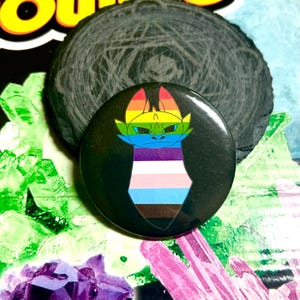May include: A black button with a cartoon dragon wearing a pride flag. The dragon is yellow, blue, and green with a rainbow mane. The pride flag is a transgender pride flag with the colors of blue, pink, white, light blue, dark blue, and brown.
