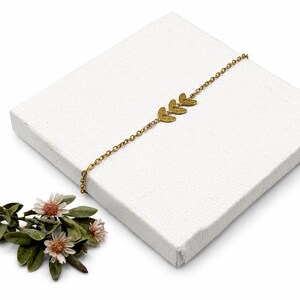 May include: A delicate gold-colored bracelet featuring three leaf-shaped charms. The bracelet is displayed on a white surface, with a sprig of flowers and green leaves in the lower left corner. The bracelet's chain is fine and elegant.