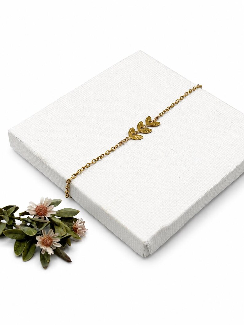 May include: A delicate gold-colored bracelet featuring three leaf-shaped charms. The bracelet is displayed on a white surface, with a sprig of flowers and green leaves in the lower left corner. The bracelet's chain is fine and elegant.
