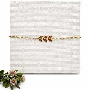 May include: A delicate gold-colored bracelet featuring a trio of leaf-shaped charms. The bracelet is displayed on a white textured surface, with a small floral arrangement in the lower left corner. The leaves are a darker gold color.