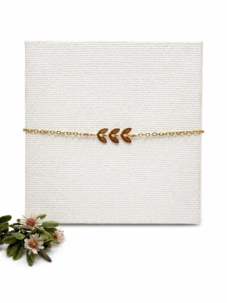 May include: A delicate gold-colored bracelet featuring a trio of leaf-shaped charms. The bracelet is displayed on a white textured surface, with a small floral arrangement in the lower left corner. The leaves are a darker gold color.