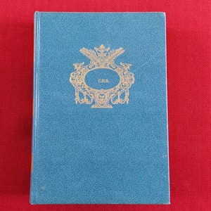 May include: A blue book with a gold embossed design on the front cover. The design features a crest with a crown and the letters "C.M.S."