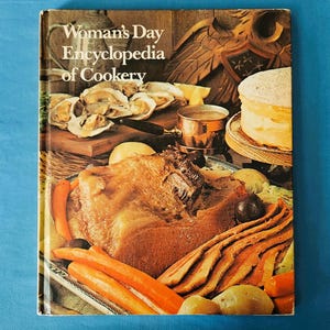 May include: A vintage cookbook titled "Woman's Day Encyclopedia of Cookery" with a cover featuring a spread of cooked food. The image includes roasted meat, carrots, potatoes, oysters, and a layered cake. The book is on a blue background.