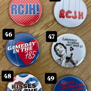 May include: A collection of round buttons with various designs and text. One button has a red and white polka dot background with "RCJH!". Another says "RCJH" in red. Other buttons say "GAMEDAY IN THE 785", "Lawrence is calling and I must go", "KISSES FOR THE HAWKS!", and "I heard the hawks are HOT".