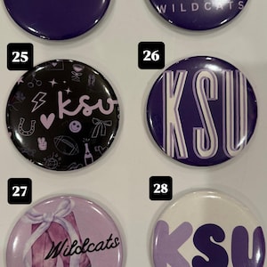 Puede incluir: Una colección de botones redondos en varios diseños y colores. Algunos botones presentan texto como "EMAW" y "KSU", mientras que otros muestran gráficos como un corazón con alas, una herradura y un balón de fútbol. Los botones son principalmente morados, negros y blancos.