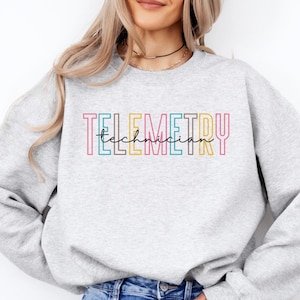 May include: A light gray sweatshirt with the word "TELEMETRY" in colorful block letters and "technician" in cursive. The sweatshirt is paired with blue jeans. The word "Ash" is written in a script font.