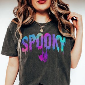 A model wears a pepper color shirt that says Spooky in an arched drippy font with a hanging bat hanging below the 2 Os. The colors are a pastel-like rainbow of iridescent colors gradient from one corner to the other. It has a retro, preppy, holographic vibe.