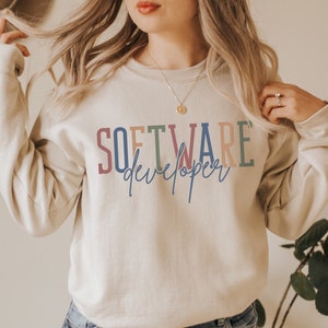 May include: A light beige sweatshirt with the text "SOFTWARE developer" printed in colorful letters. The text is in a rainbow color scheme with a blue, green, pink, and orange color palette.