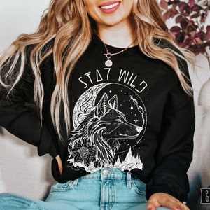 A model wears a black sweatshirt that says Stay Wild in a futuristic arch and a boho graphic in white outline of a profile of Wolf in front of a crescent moon with stars. There are trees in the distance. The wolf has some flowers on their neck.