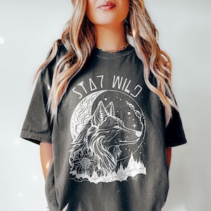 A model wearing a pepper colored shsirt with an outline graphic of a wolf with a crescent moon behind it and some flowers and trees beneath. The words Stay Wild in a futuristic font are written above.