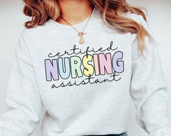 Op de afbeelding: Een lichtgrijs sweatshirt met de tekst "certified nursing assistant" in regenboogkleuren. De tekst is in een speels, handschriftlijk lettertype.