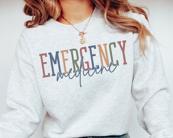 May include: A light gray sweatshirt with the text "EMERGENCY medicine" printed in colorful letters. The text is in a rainbow color scheme with shades of red, orange, yellow, green, blue, and purple.