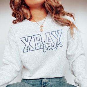 A model wears an ash colored sweatshirt that says Xray tech. Xray is written in a large blocky outline font in different shades of blue from dark to light-gray-blue. Tech is written in smaller lowercase script in the lower right of xray.