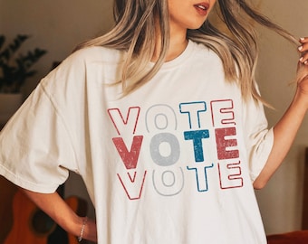 May include: A white oversized t-shirt with the word "VOTE" printed in red, blue, and gray letters. The shirt is worn by a person and has a distressed vintage look. The color of the shirt is ivory.