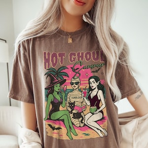 A model wears an espresso color shirt with a retro summerween graphic of a witch, a mummy, and a woman with elf pointy ears sitting on a beach with a black cat in front os a sunset and a palm tree. It says Hot Ghouls summer above them with some bats flying around.