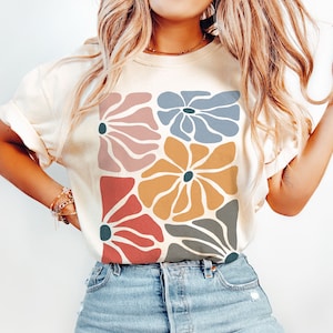 A model wears an ivory shirt with abstract, wavy, boho flowers in colors of muted mauve, light grey-blue, gold, red, and dark taupe all with deep blue pistils. The colorful design is a slightly distressed texture.