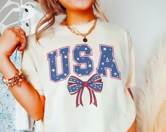May include: A white t-shirt with a blue and red bow and the word "USA" in blue with white stars. The shirt is on a person wearing blue jeans. The text "Ivory" is in the bottom right corner of the image.